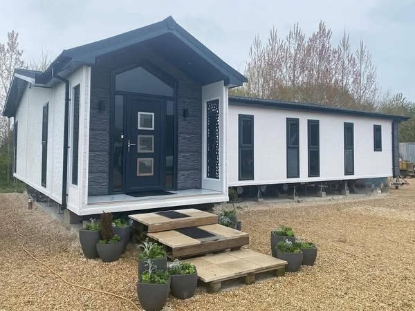 Modern prefab home with a gabled roof, dark door, and multiple tall windows. Surrounded by gravel and potted plants, creating a serene setting.