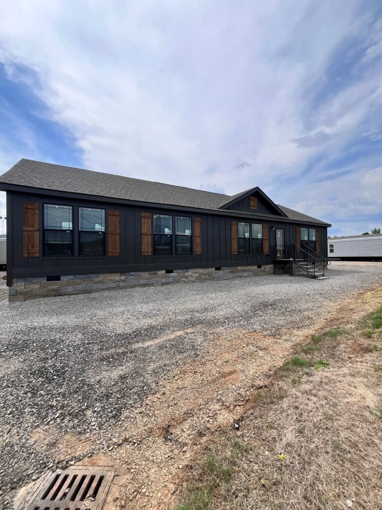 A modern, dark-colored manufactured home with wooden shutters and large windows sits on a gravel driveway. A cloudy sky looms overhead.
