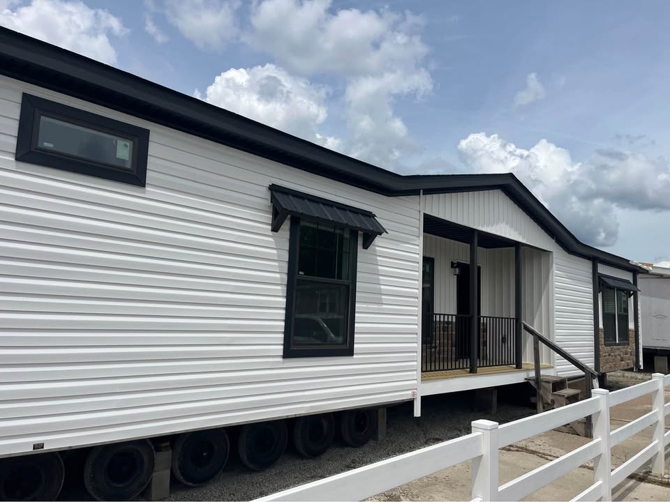 White mobile home with black-trimmed windows, front porch, and stairs. Overcast sky and a white fence in the foreground; calm, suburban setting.
