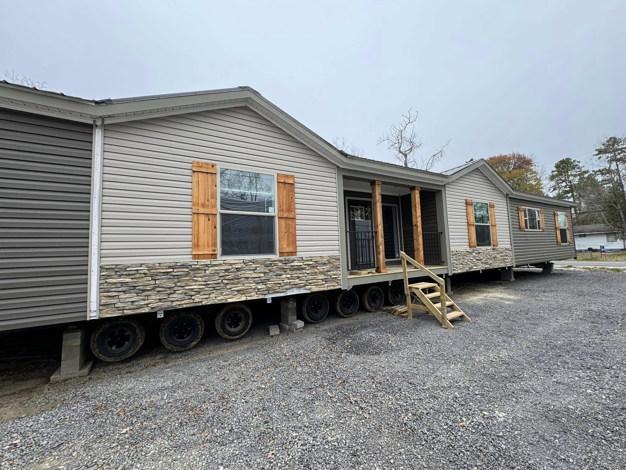 Single-story manufactured home with stone facade and wooden shutters, elevated on wheels in a gravel lot. Overcast sky, sparse trees in background.