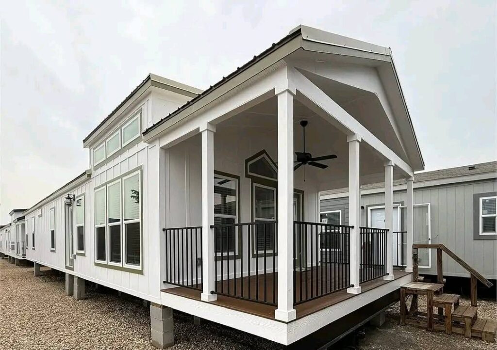Modern mobile home with a white exterior and a small porch featuring a ceiling fan and black railings. Several large windows line the side. Gravel pathway in front.