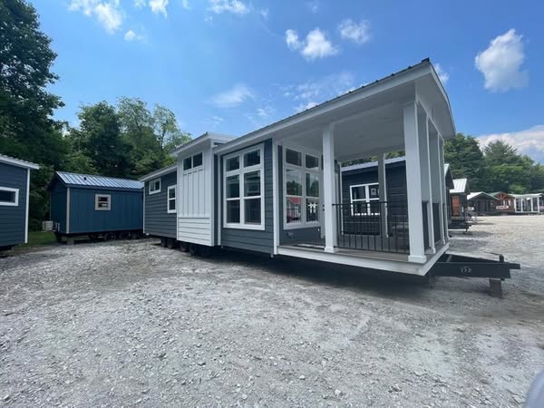 Small gray and white modular home with large windows and a spacious porch. Set on a gravel lot with trees and a clear sky in the background.