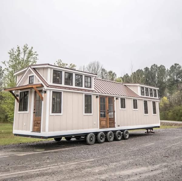 Tiny house on wheels wTiny house on wheels with cream siding and numerous windows sits on a gravel path surrounded by trees. The atmosphere is serene and rustic.ith cream siding and numerous windows sits on a gravel path surrounded by trees. The atmosphere is serene and rustic.