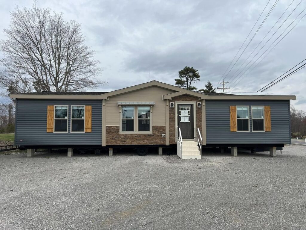 A manufactured home with a central beige section and blue side wings. It has a white front door, matching steps, and yellow window shutters. Overcast sky.