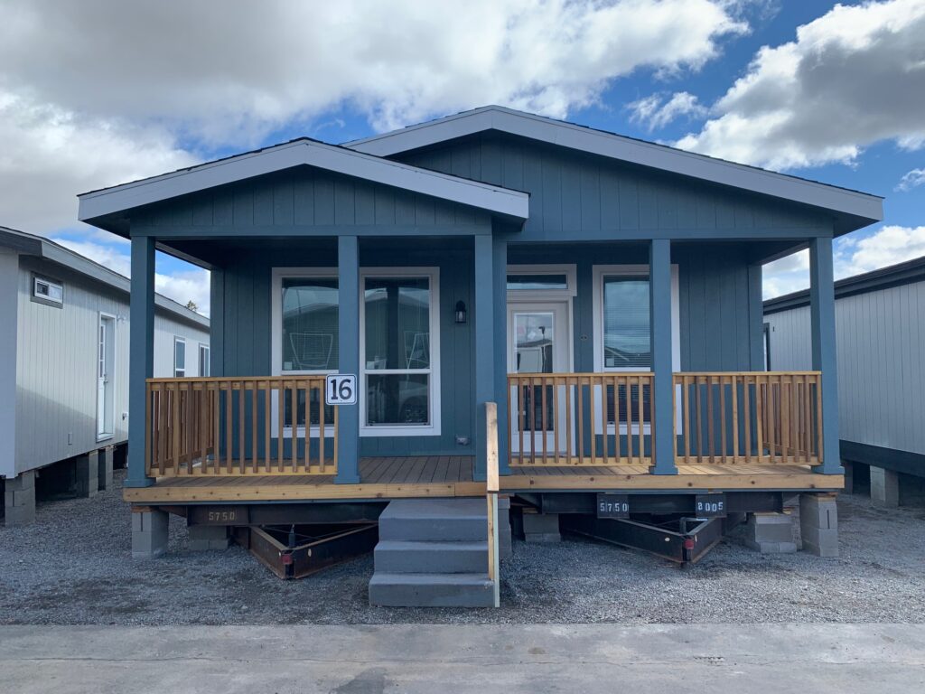 A blue mobile home with a wooden porch and railing stands on a gravel lot under a partly cloudy sky. The number 16 is displayed on the porch.