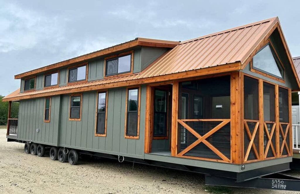 Modern tiny house on wheels with green siding and a copper metal roof. Features multiple windows and a screened porch, set on a gravel surface.