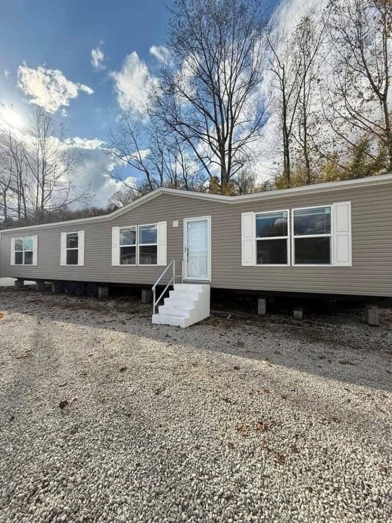 A beige modular home with white shutters and a central white door sits on a gravel lot. Leafless trees surround the house under a partly cloudy sky.