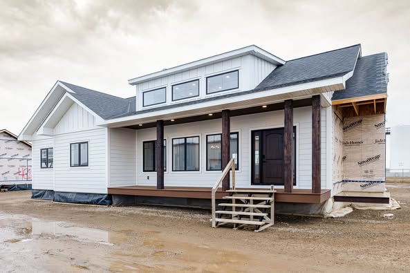 A modern, partly constructed house with white siding and a dark roof sits on a muddy plot. It features a front porch with black columns and multiple windows, conveying a cozy yet unrefined atmosphere.