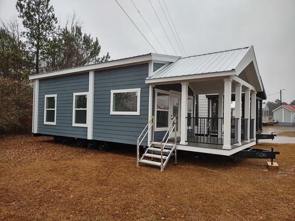 A modern tiny house on wheels with blue siding, white trim, and a small porch stands on a grassy area under a gray, overcast sky.