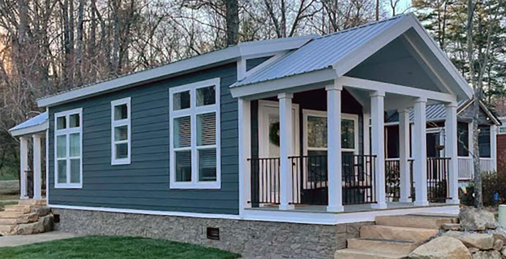 A charming tiny house with blue siding and white trim sits surrounded by trees. It features large windows and a welcoming porch, creating a cozy atmosphere.