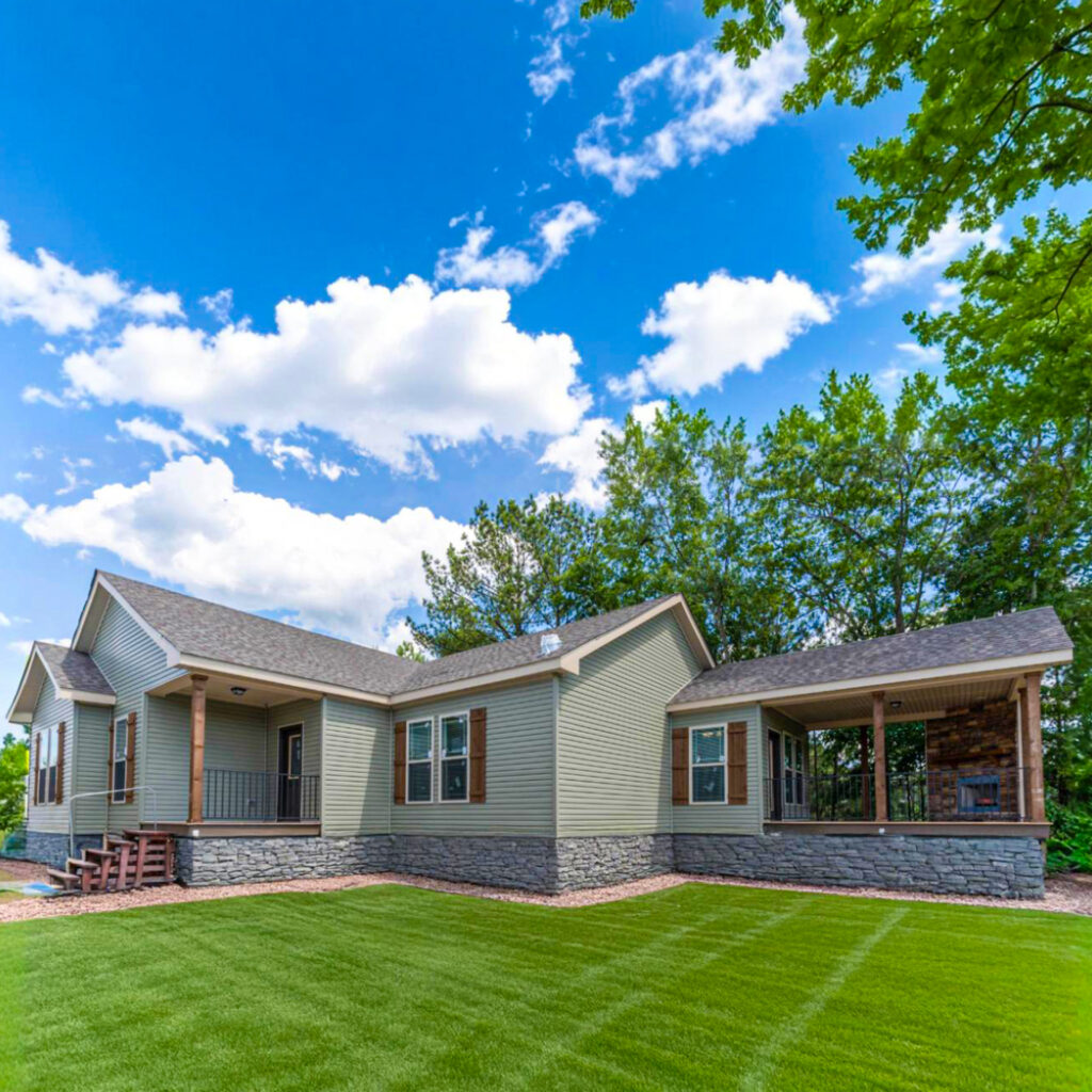 A single-story house with gray siding and a stone foundation sits on a lush, green lawn. The sky above is blue with fluffy white clouds, evoking a serene atmosphere.