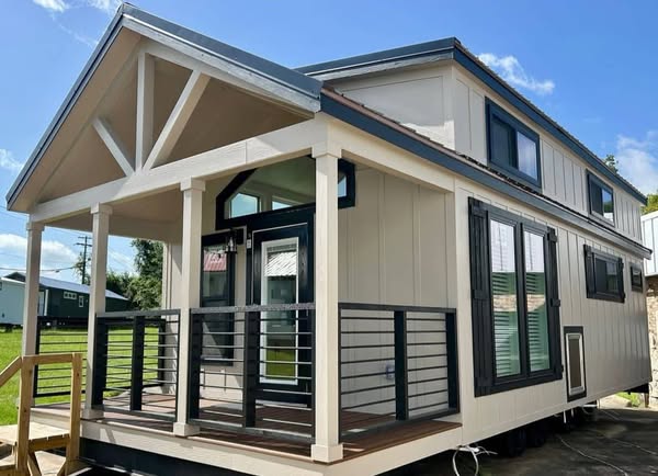 A modern tiny house with a beige exterior and black trim. It features a covered porch, large windows, and a sloped roof, set against a clear blue sky.