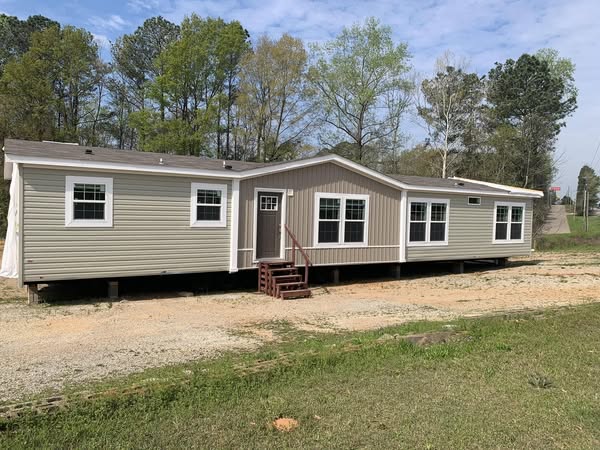 Manufactured home with beige siding and white trim, set on a grassy area with steps leading to the door. Trees and a blue sky in the background.