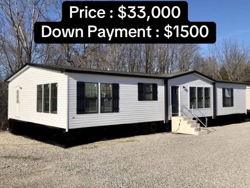 White mobile home on a gravel lot, surrounded by leafless trees. Text above states: “Price: $33,000, Down Payment: $1,500.” Bright, clear day.