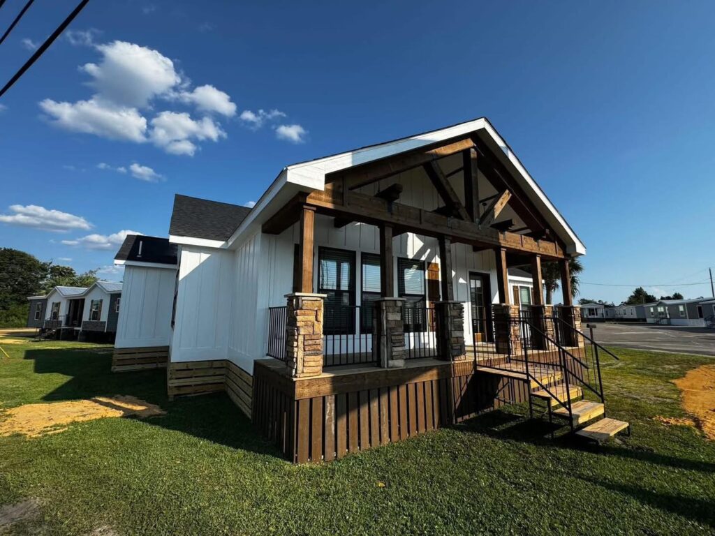 A small, modern white house with a wooden porch and stone accents stands under a bright blue sky. The setting is calm, with lush green grass and a few clouds.