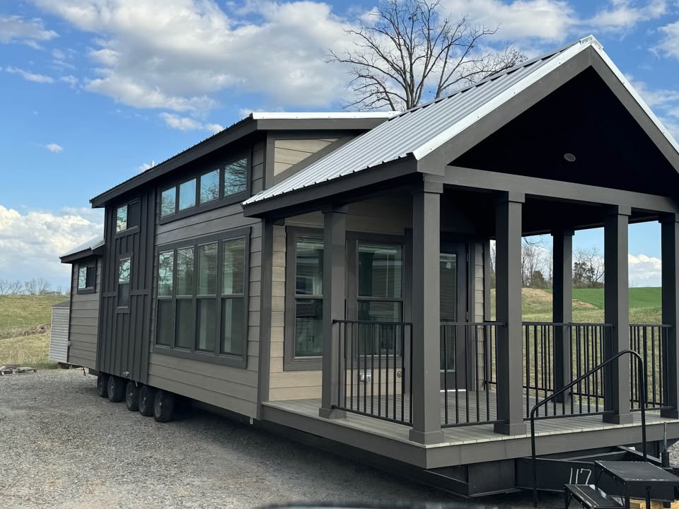 Modern tiny house on wheels with a gabled roof and large windows, situated on gravel. The porch has railings, set against a backdrop of grass and a blue sky.