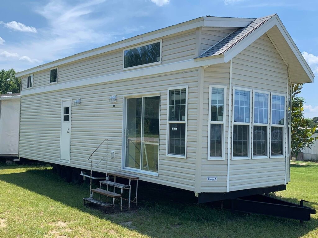 A beige tiny house on wheels is parked on grass under a clear blue sky. It features large windows, a sliding glass door, and a small metal staircase.