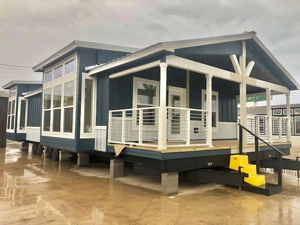 Modern modular home with large windows and a small porch, painted in dark blue with white trim. Wet pavement and overcast sky suggest recent rain.