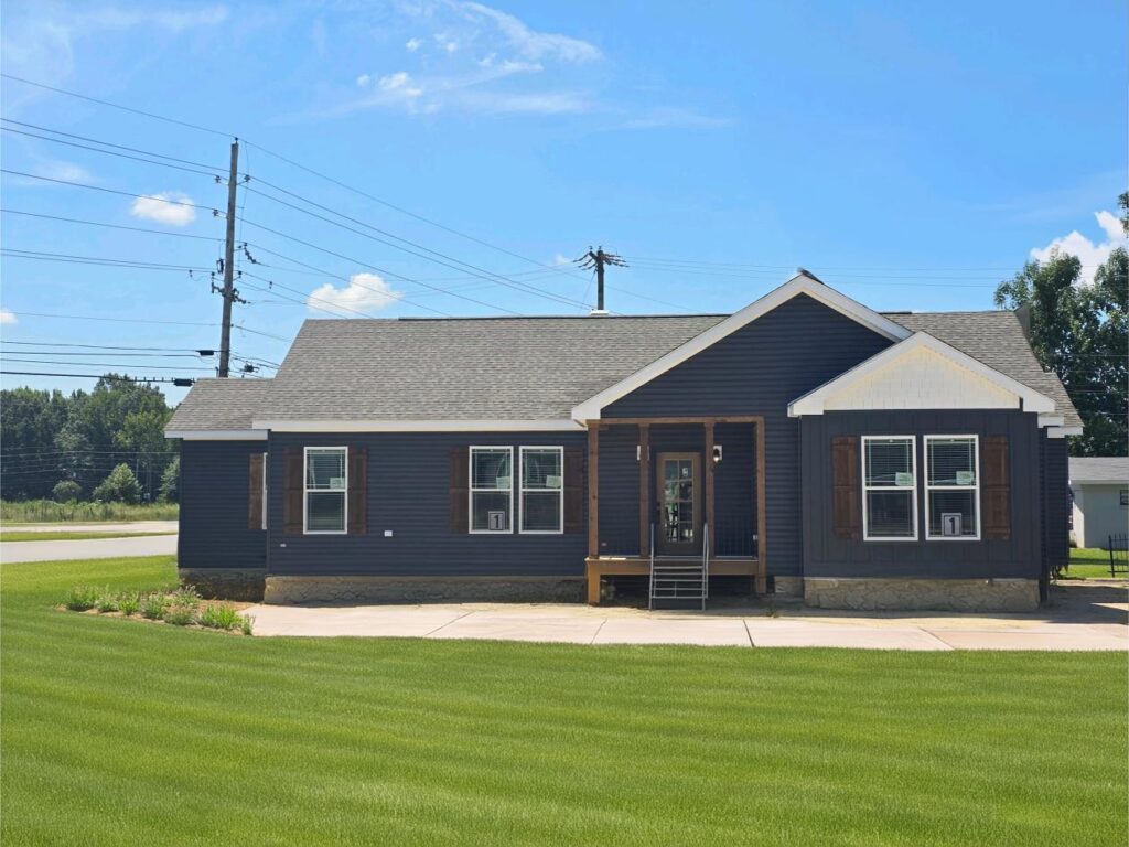 Single-story dark blue house with white trim and wooden shutters, set against a bright blue sky. The yard is well-maintained with green grass.