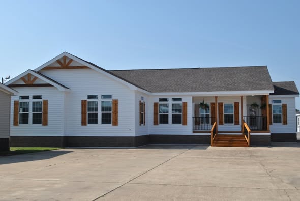 A white single-story house with a grey roof features large windows with wooden shutters, a small porch, and wooden steps, set against a clear blue sky.