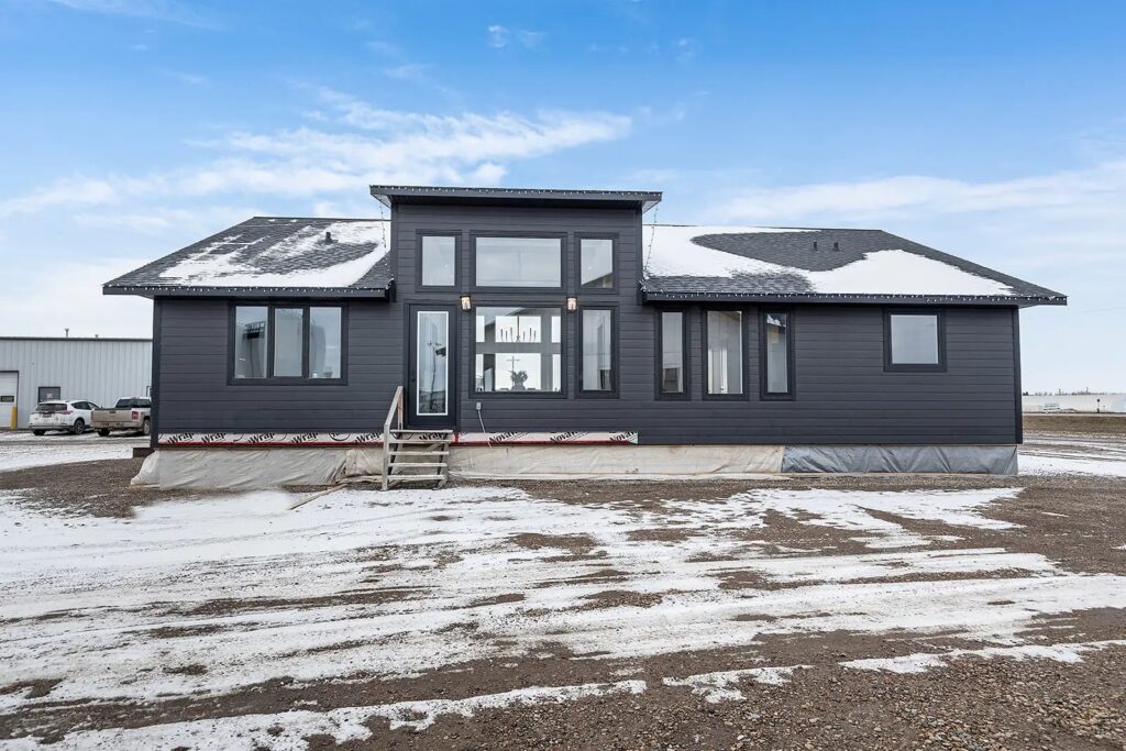 Modern grey house with large windows and snow on the roof, set against a bright blue sky. The snowy yard adds a serene, wintry feel.