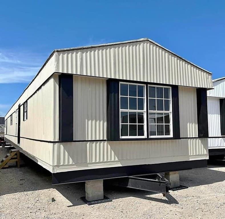 Beige mobile home with dark accents stands on concrete blocks under a bright blue sky. It features large front windows, creating a simple and stable appearance.