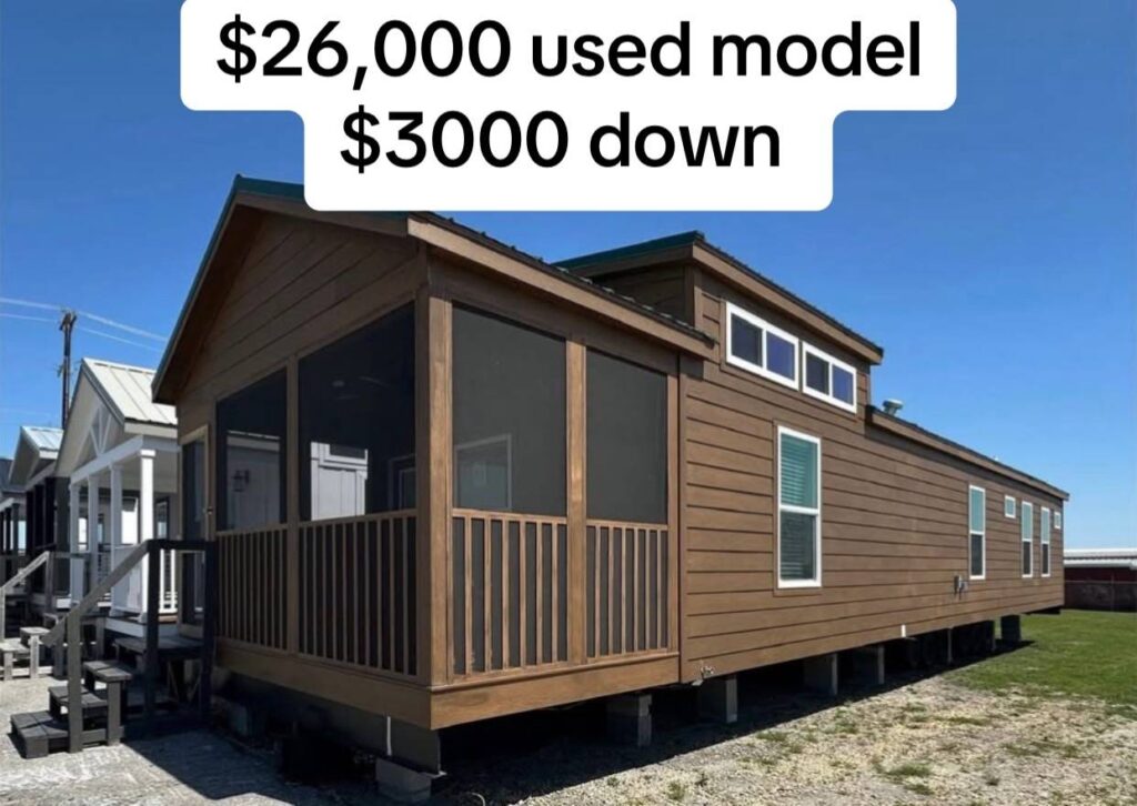 Brown manufactured home with a screened porch on a sunny day. Price above reads "$26,000 used model, $3000 down." Other homes visible in the background.