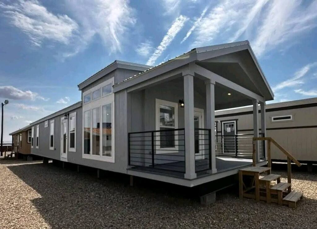 A modern gray mobile home with a covered porch and stairs sits on a gravel lot under a partly cloudy sky, conveying a sense of simplicity and comfort.