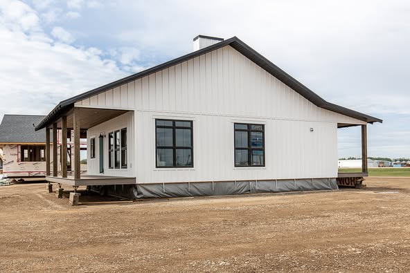 A modern, white single-story house sits on a dirt lot under a cloudy sky. Large windows and a wrap-around porch suggest openness and relaxation.