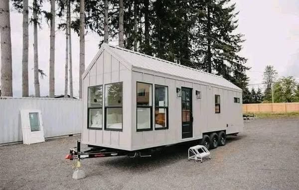 A modern tiny house on wheels with large windows, set against a backdrop of tall trees. The minimalist design conveys a sense of cozy, mobile living.