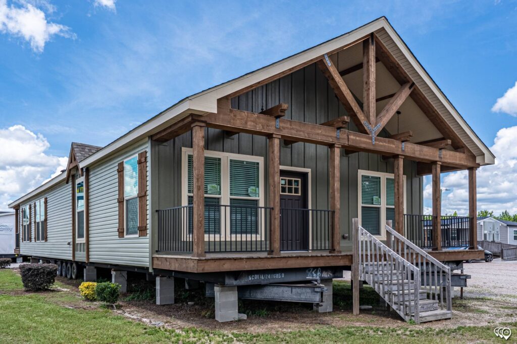 A modern manufactured home with a rustic wooden porch under a clear blue sky. The home rests on stilts with steps leading to the front door.