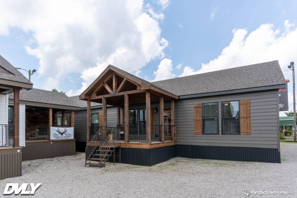 A modern manufactured home with a peaked porch roof and wooden accents sits on gravel. The sky is partly cloudy, adding a serene backdrop.