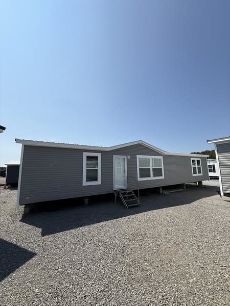A light grey mobile home with a white door and windows sits on a gravel lot under a clear blue sky. The scene is calm and minimalist.