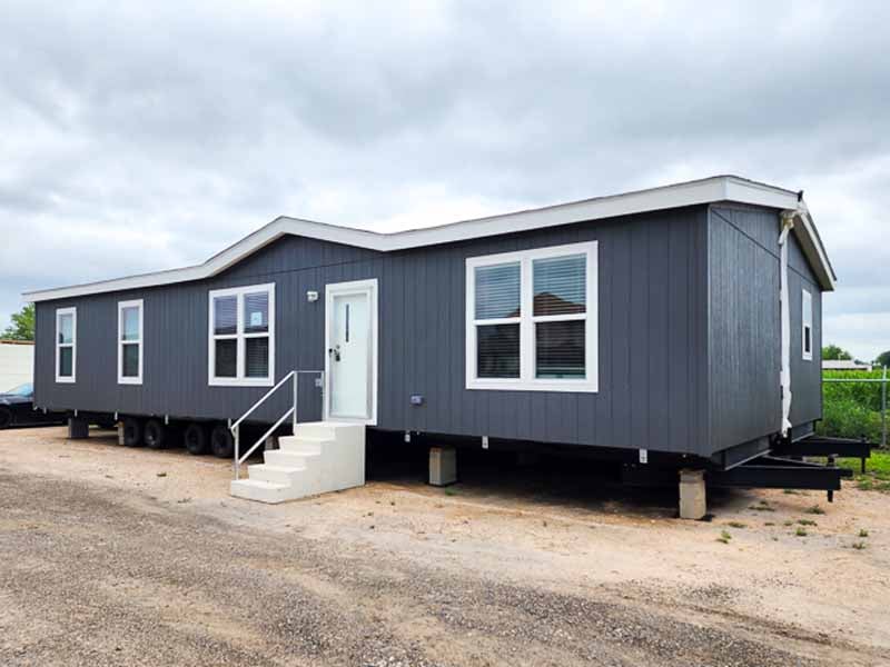 Gray modular home on wheels, with a white door and large windows, placed on a gravel lot under a cloudy sky. Steps lead to the entrance, conveying a simple, functional design.