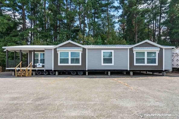 A gray manufactured home with a metal roof is elevated on wheels. Large windows highlight its modern design. Tall pine trees set a tranquil backdrop.