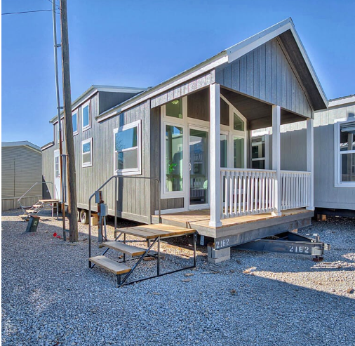 Compact modern tiny house with large windows and a small porch, set on gravel. Bright blue sky enhances its clean, minimalist appearance.
