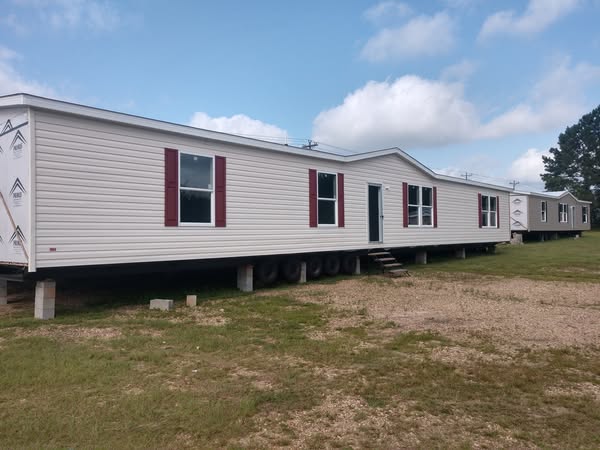 A white mobile home with red shutters is set on a grassy field under a cloudy sky, elevated on blocks. It has wheels and a small wooden staircase.