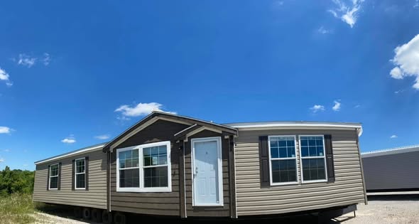 A tan mobile home with a white door and multiple windows stands under a clear blue sky, conveying a sense of openness and tranquility.