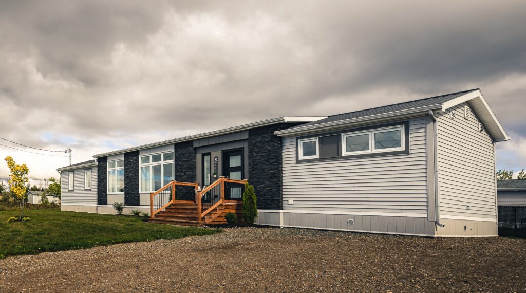 Modern manufactured home under a cloudy sky, featuring a dark facade with large windows, wooden steps, and a gravel driveway, creating a serene mood.