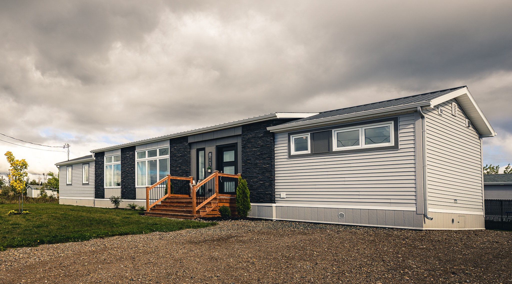 Modern manufactured home under a cloudy sky, featuring a dark facade with large windows, wooden steps, and a gravel driveway, creating a serene mood.