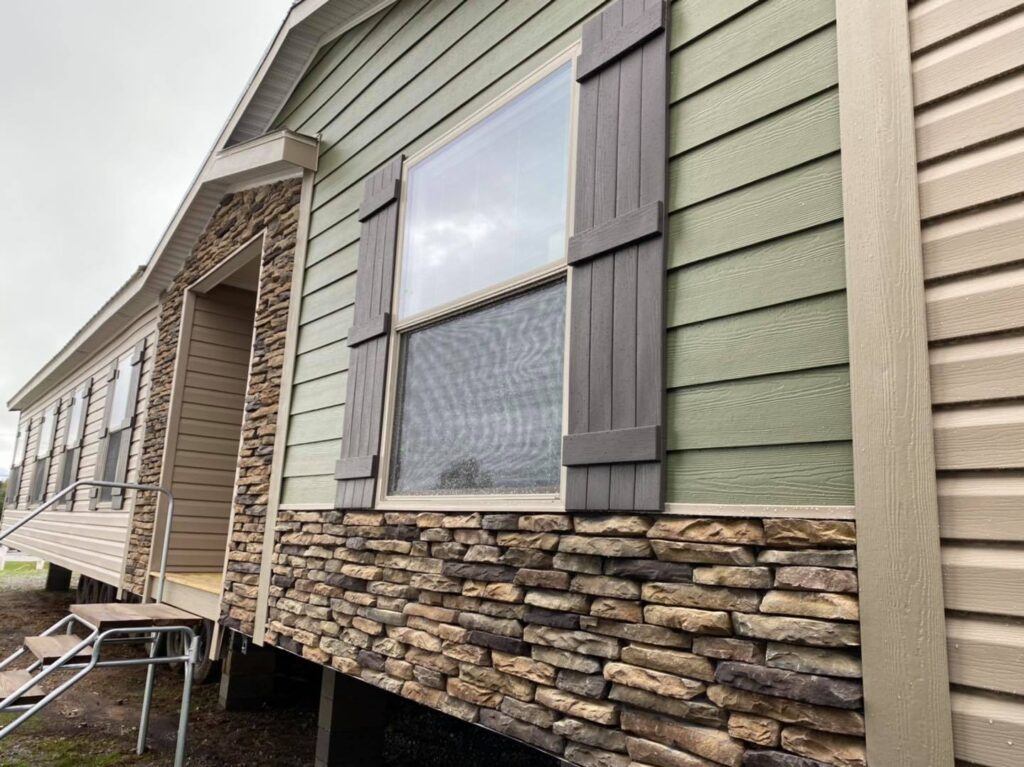 Side view of a manufactured home with stone and beige siding. Large window with shutters, metal steps leading to a porch, and an overcast sky.