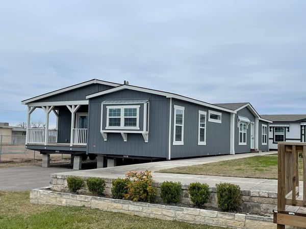 A gray manufactured home with a small porch and white trim sits on a lot. A stone-bordered lawn with shrubs and cloudy skies complete the cozy scene.