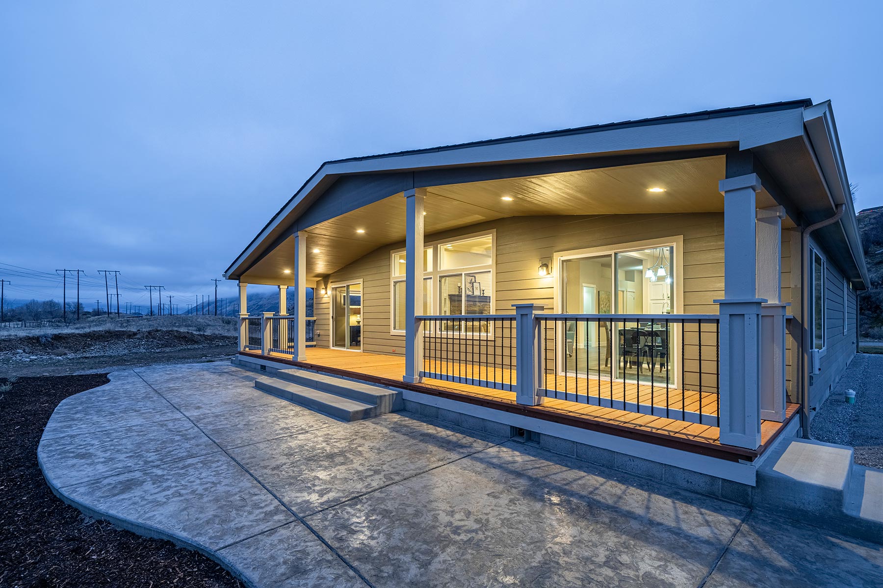 Modern single-story house at dusk, featuring a well-lit, spacious porch with wooden railing. The sky is overcast, creating a cozy atmosphere.