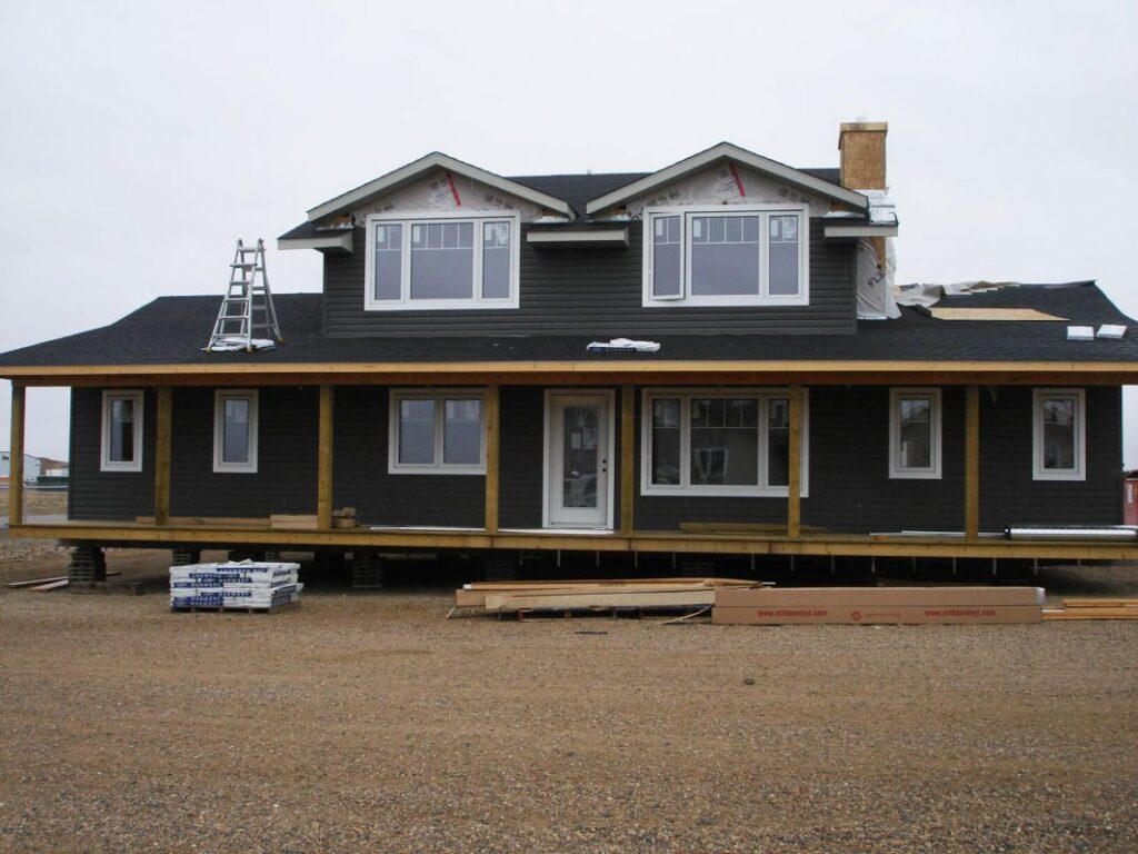 Two-story dark gray house under construction with white trim. Ladders and building materials lie around the gravel lot, creating a work-in-progress feel.