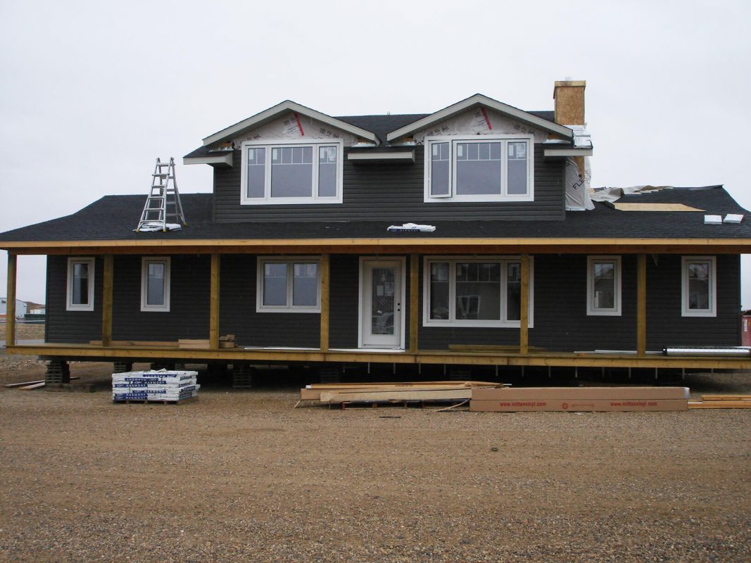 Two-story dark gray house under construction with white trim. Ladders and building materials lie around the gravel lot, creating a work-in-progress feel.