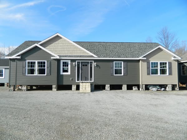 Single-story modular home on a gravel lot, featuring gray siding, white trim, and large front windows under a clear blue sky.