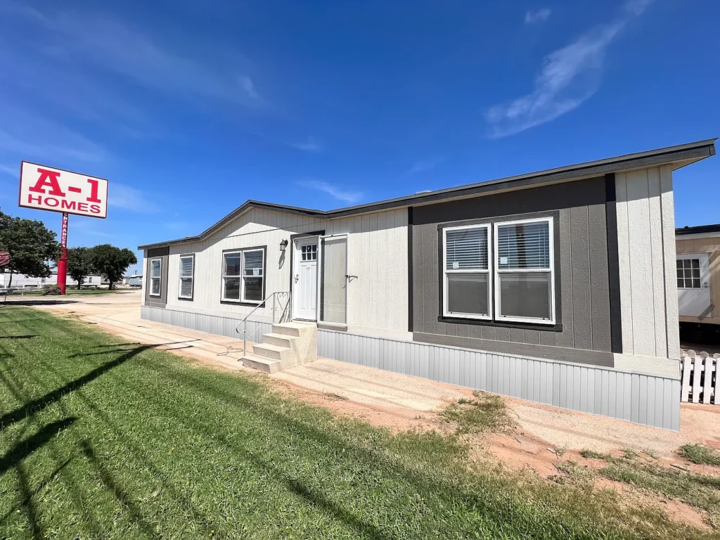 Single-story manufactured house with gray and white paneling and multiple windows, set on a grassy lot. An "A-1 Homes" sign is visible in the background under a clear blue sky.