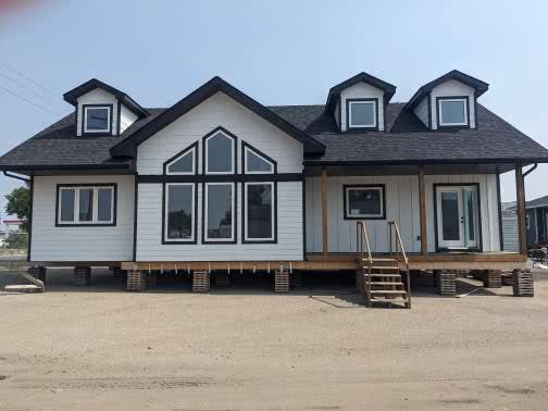 A white, modern house with black trim sits elevated on blocks. It features large triangular windows, three dormers, and a small wooden staircase.