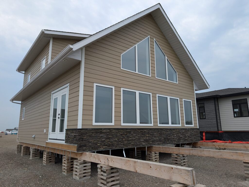 A beige two-story house with large windows and stone accents is elevated on wooden stilts. The setting is overcast, conveying an unfinished construction site.