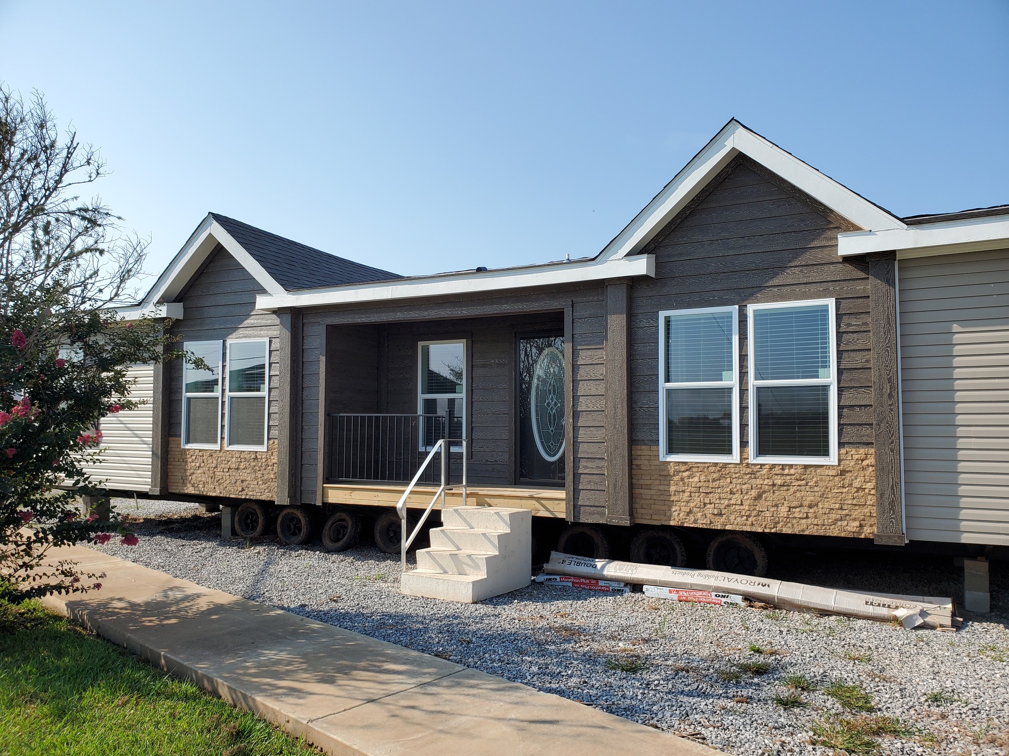 Single-story modular home with dark siding and white trim, featuring three peaked roofs. A small porch and stairs lead to an oval glass door. Bright, clear day.